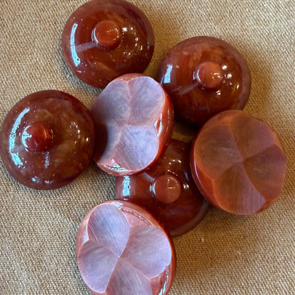 Six polished reddish-brown buttons arranged on a beige fabric background, with two buttons showing their floral faceted sides and four showing their domed tops.