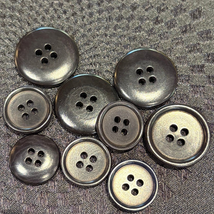 A group of nine black buttons with four holes, arranged on a textured black fabric background.