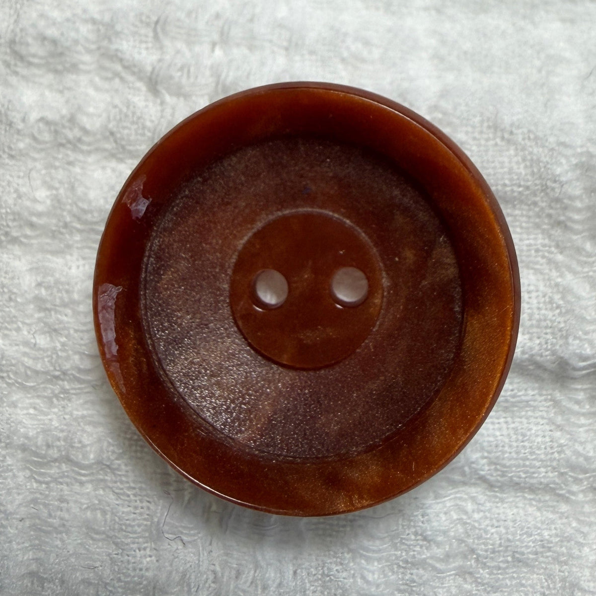 A single round brown button with two holes, photographed from above on a textured white fabric background.