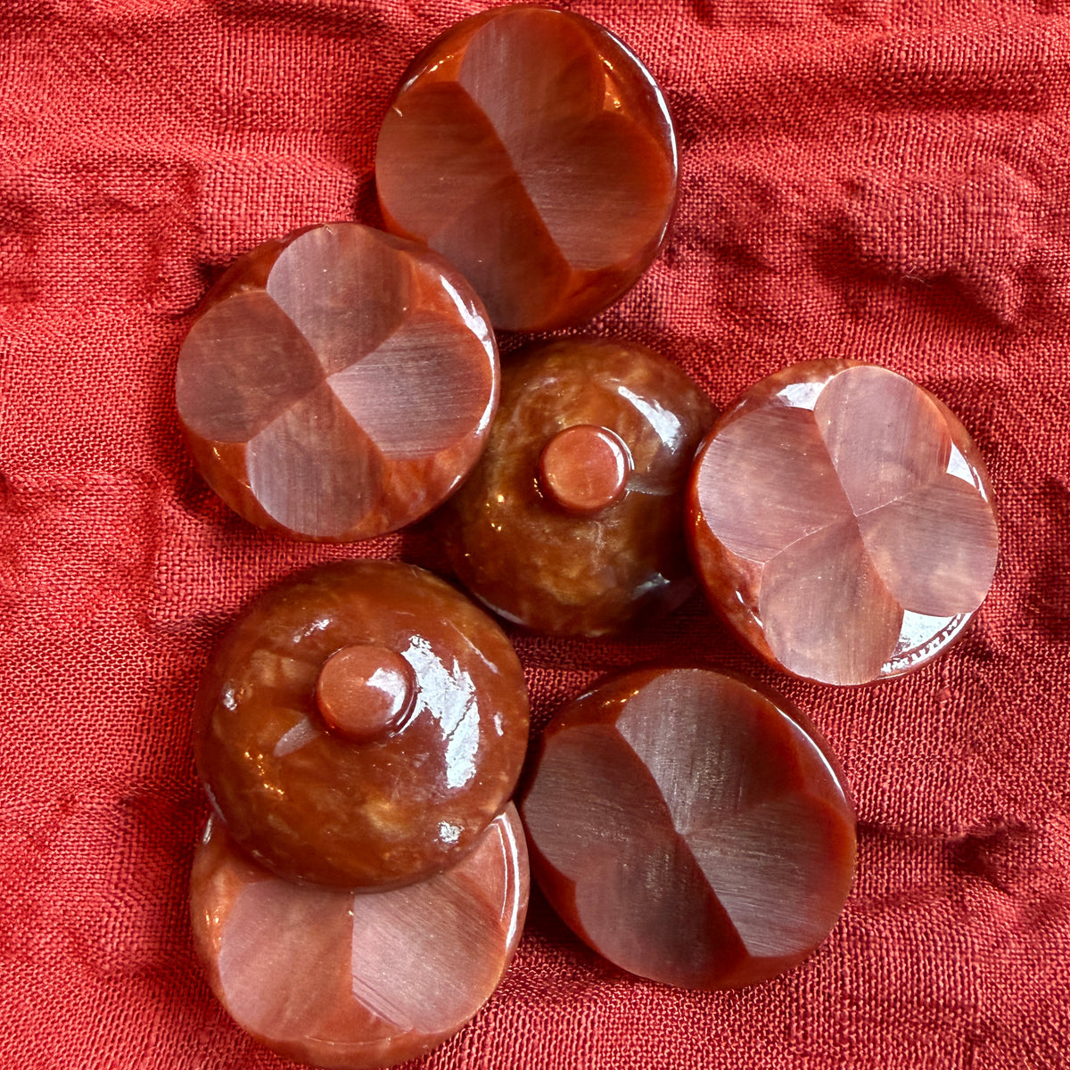 Six round carved buttons in shades of brown and reddish-brown, arranged on a textured red fabric background.