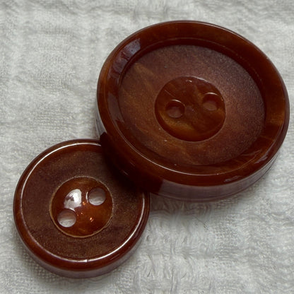 Two round amber-brown buttons with two holes, one larger and one smaller, placed on a textured white fabric background.